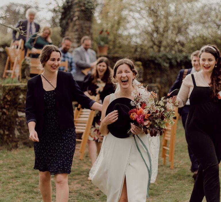 Laughing bride in Charlie Brear wedding dress and daisy applique veil holds black fedora and multicoloured bridal bouquet whilst surrounded by smiling wedding guests at garden party wedding in Devon