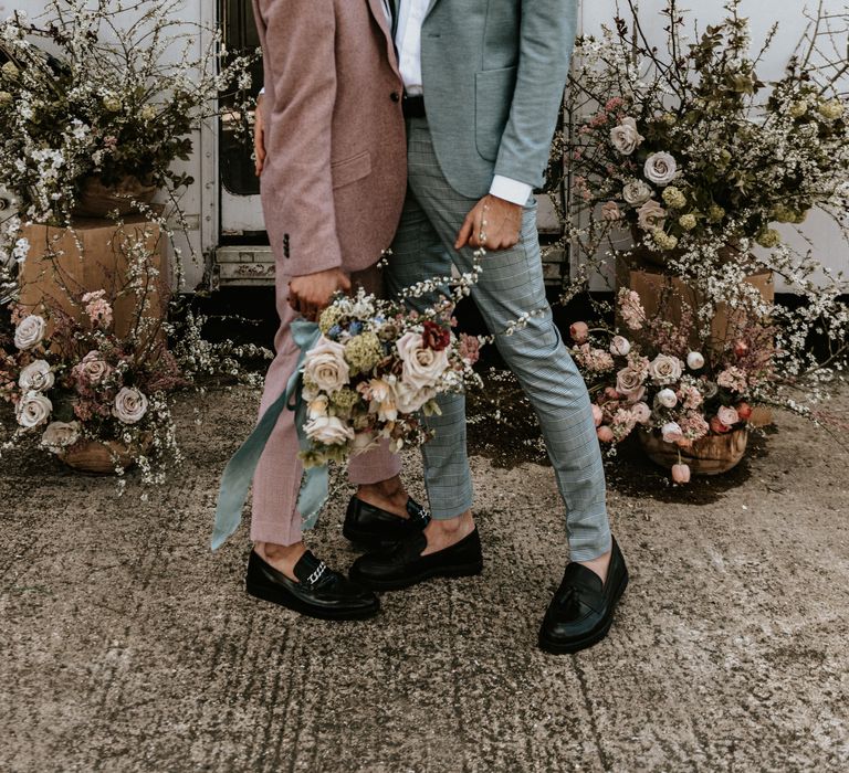 Grooms smile and laugh in front of rustic floral installations whilst holding floral bouquet 