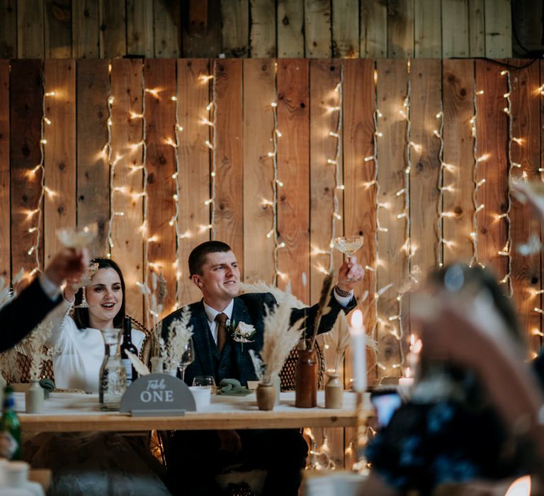 Bride and groom raising a glass sitting at their sweetheart table with a fairy light backdrop 