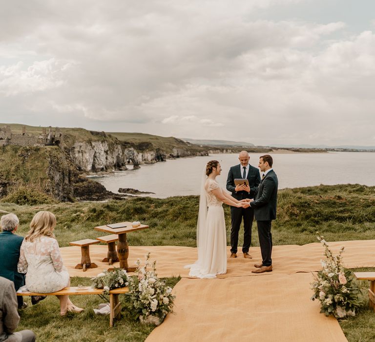 Smiling bride in lace top with capped sleeves and satin skirt holds hands with groom in navy suit at clifftop ceremony for Dunluce Castle wedding