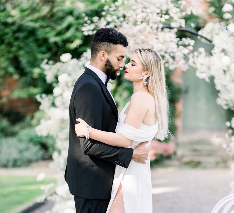 Bride in an off the shoulder wedding dress with front split embracing her husband in a black tie suit