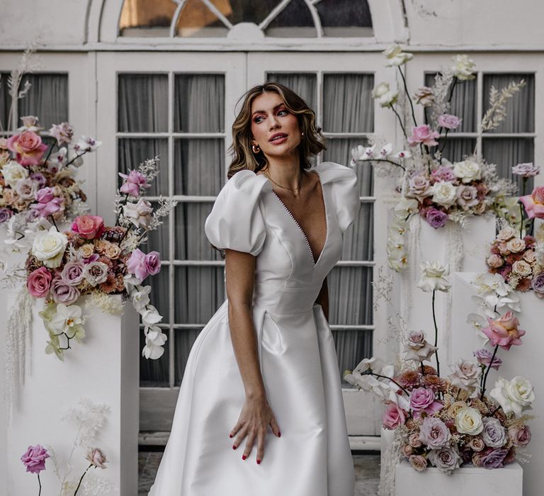 Stylish bride in a Love In Lace bridal gown with puff sleeves and plunging neckline surrounded by pink wedding flowers 