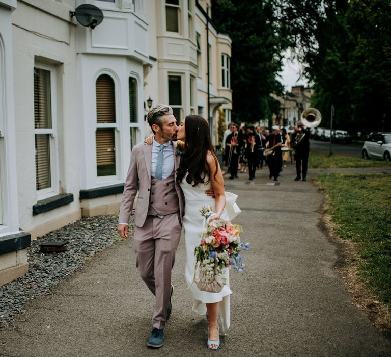 Bride in white strapless Rebecca Vallance Dress carrying ivory bridal bag and multicoloured bridal bouquet kisses groom in brown Moss Bros suit and blue tie as they walk through streets of Harrogate followed by the New York Brass Band