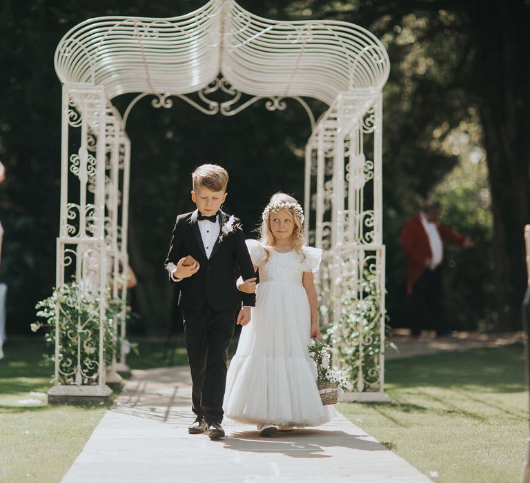 A young girl in a white dress walks down a wedding aisle with a young boy in a black tuxedo. The aisle is outside.