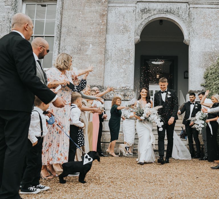 Bride in white Pronovias wedding dress holding white, pink and green wedding bouquet walks out of Came House Dorset with groom in black tuxedo as guests throw confetti