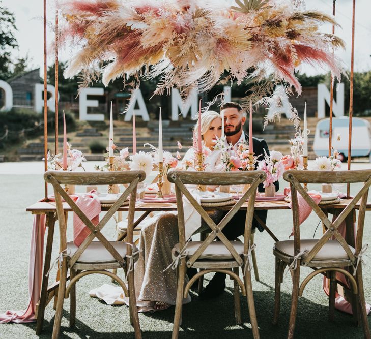 Bride and groom sitting at their outdoor reception at Dreamland Margate with pink coloured and natural pampas grass installation, and pink and gold wedding table decor 