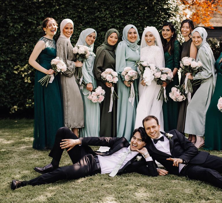 Bride with all her bridesmaids in various hades of green and style of dress with playful groomsmen laying on the grass in front