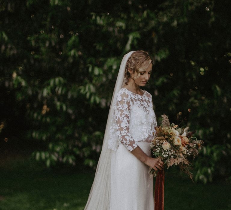 Bride in white Rime Arodaky wedding dress and mesh embroidered floral top with three-quarter length sleeves stands wearing church length veil and holding mixed burnt orange bridal bouquet at garden wedding with burnt orange wedding theme