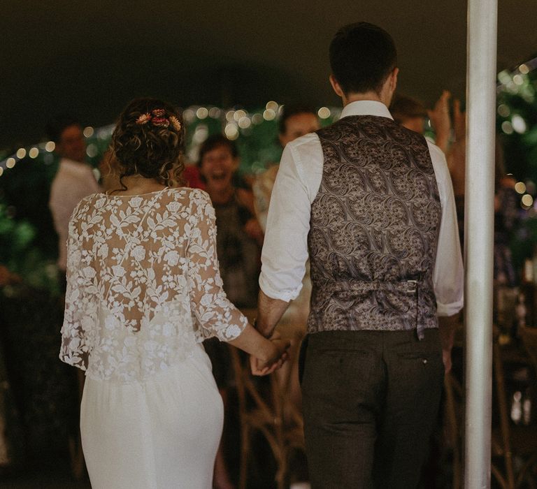 Bride in white Rime Arodaky dress and mesh floral embroidered top walks into tipi wedding reception holding hands with groom in patterned back waistcoat and white shirt for garden wedding with burnt orange wedding theme