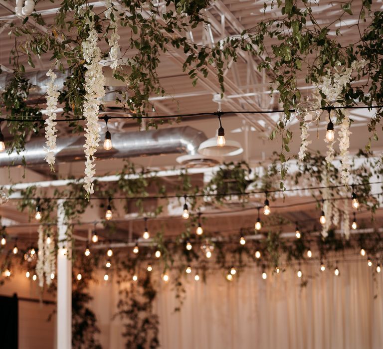 White frosted cake sits within wedding venue on a table covered in a white cloth, whilst hanging florals can be seen from above