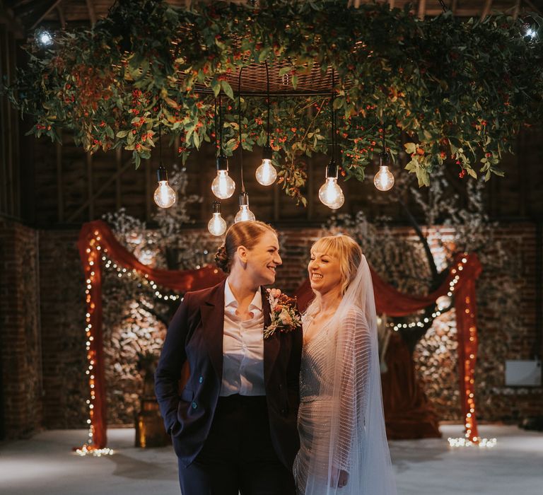 Two brides smiling, standing under Edison bulbs and hanging foliage at pumpkin spice wedding