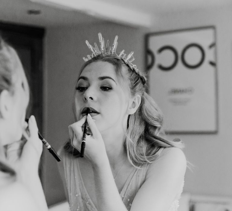 Bride in crown and pearl wedding dress does her lipstick in the mirror before barn wedding ceremony