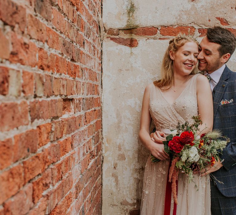 groom in a grey check suit embracing his bride in a tulle and pearl wedding dress as she holds a red wedding bouquet 