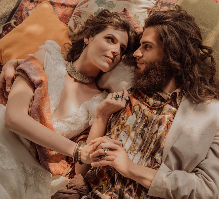 Bride & groom lay down with one another on Moroccan style rugs and neutral toned pillows as they look lovingly at one another 