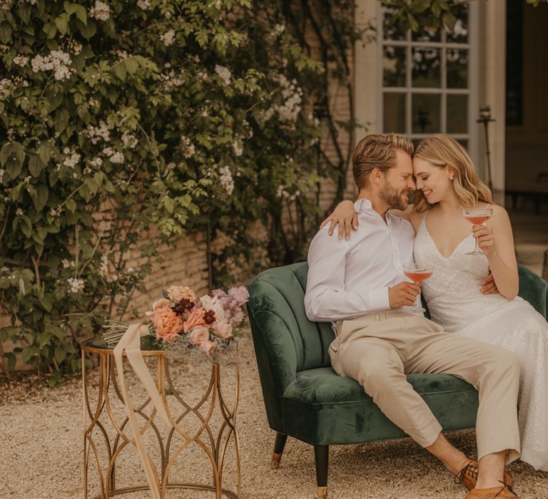 Bride and groom sitting on a green velvet sofa sipping cocktails at their outdoor wedding 