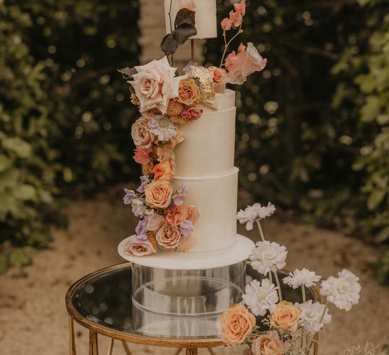 Tall three-tier buttercream wedding cake with elevated layer decorated with pastel orange, peach, coral and pink wedding flowers 