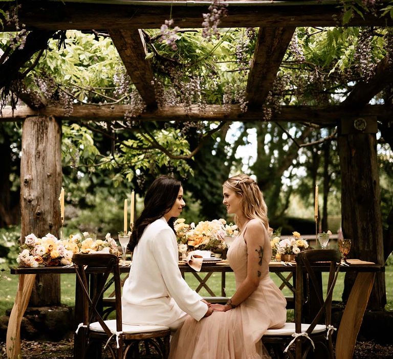 Bride in a white suit and blush pink wedding dress sitting at their al fresco wedding reception table under a pergola 