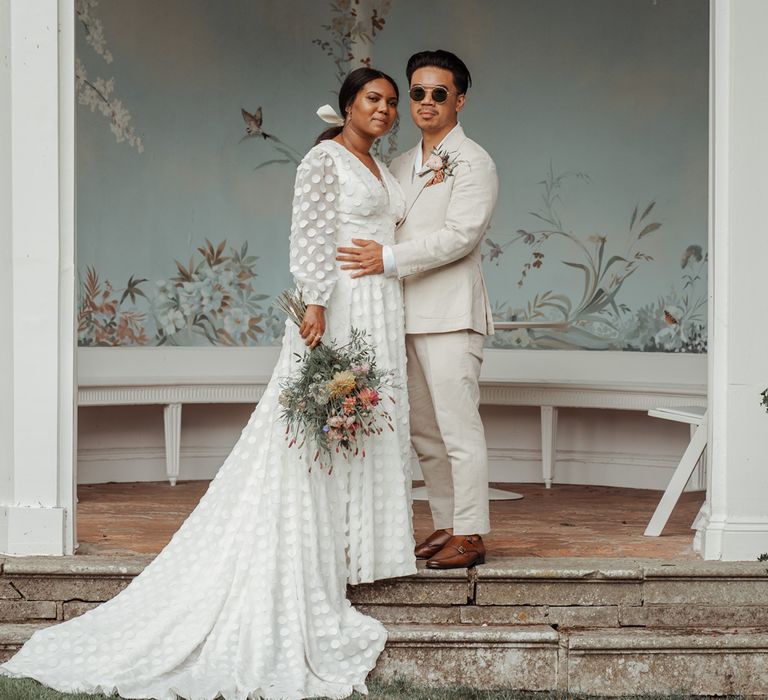 Bride in white long mesh sleeve spotted Vagabond wedding dress with train and bridal hair bow holding mixed bridal bouquet stands with groom in white shawl collared shirt and double breasted linen suit wearing sunglasses in Victorian summerhouse at Wasing Park wedding