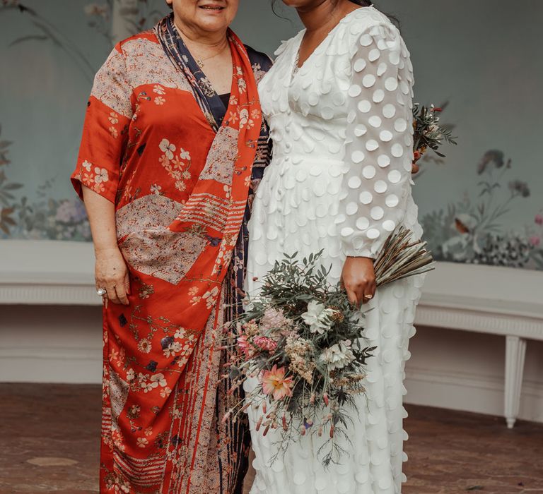 Bride in white long mesh sleeve spotted Vagabond wedding dress holding mixed bridal bouquet stands with woman in red, white and black silk dress outside Victorian summerhouse at Wasing Park wedding