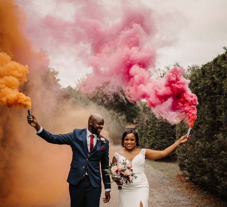 Smoke bomb wedding photo with a Black bride in fitted wedding dress with front split holding a pink grenade and a Black groom in a navy suit holding an orange flare