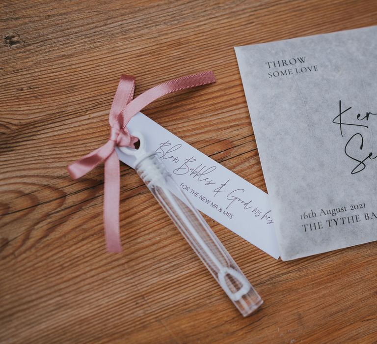 Grey paper personalised confetti bag next to bubbles tied with pink ribbon at Tythe Barn wedding with barn wedding flowers