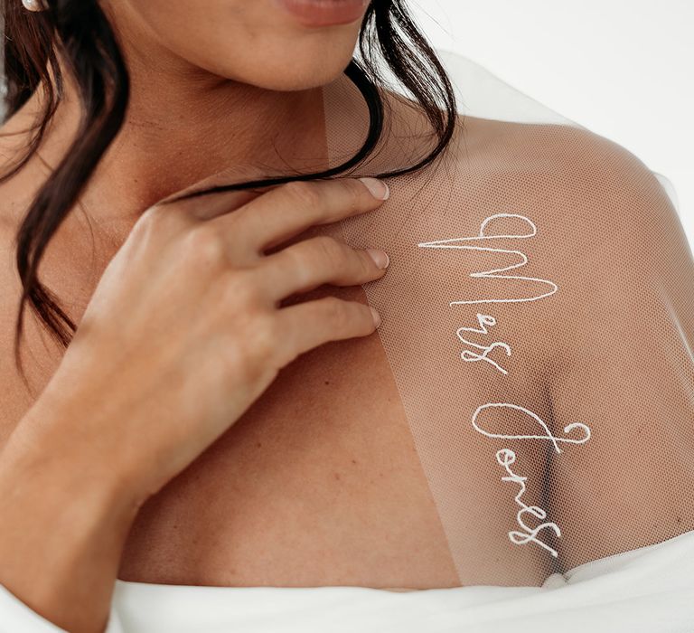 Bride in a strapless wedding dress with loosely pinned wedding hair holding her embroidered personalised veil on her shoulder to show the detail 