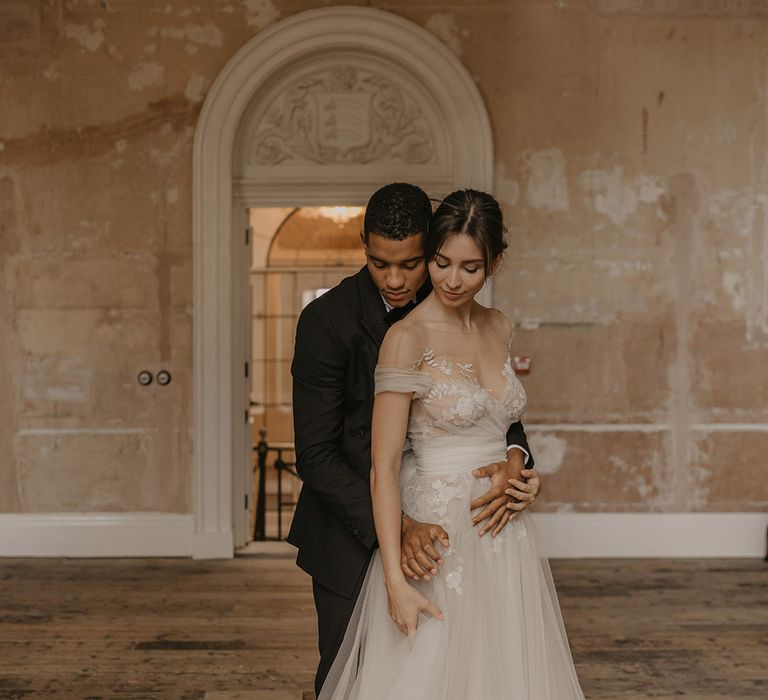 Groom in a black tie suit embracing his bride in a tulle and lace wedding dress with full skirt, Bardot off the shoulder detail and illusion neckline 