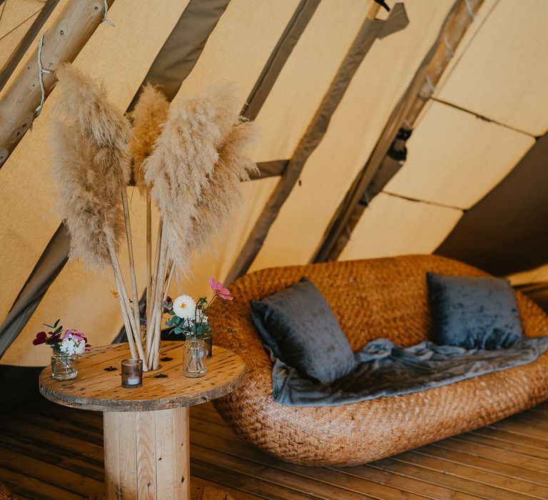 Rattan sofa with blue crushed velvet cushions and round wooden table with pampas grass and florals inside tipi for late summer wedding at Wellington Wood Norfolk