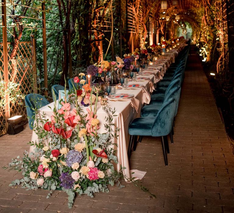 Reception table underneath pergola surrounded by trees complete with colourful tablescape, hanging lanterns and emerald blue chairs