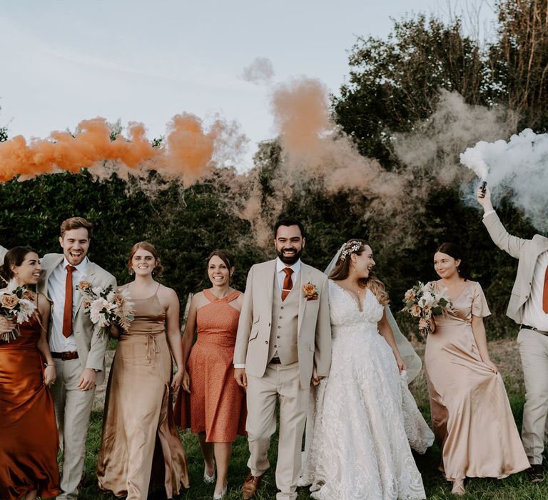 Bride in homemade lace wedding dress holds hands with groom in beige three piece suit as they walk with groomsmen and bridesmaids through field whilst holding orange and white smoke bombs