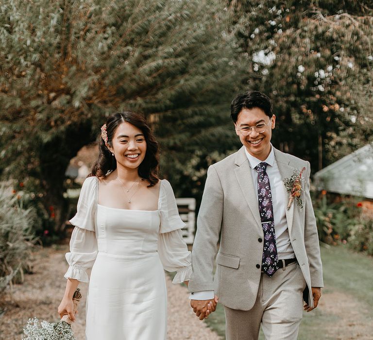 Bride & groom walk hand in hand down pathway surrounded by gardens