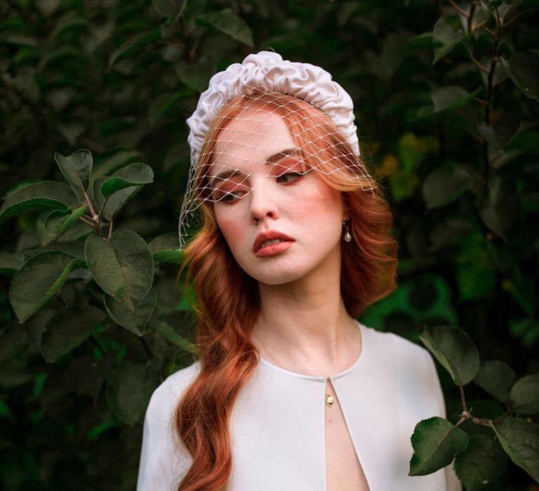 Bride in white wedding dress and bridal cape wearing white headband with net stands in orchard holding apple before marquee tent wedding reception