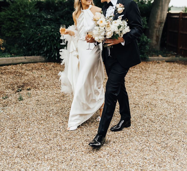 Bride in halterneck Halfpenny London wedding dress walks with groom in black tie holding brides white and peach bouquet as they both hold champagne flutes