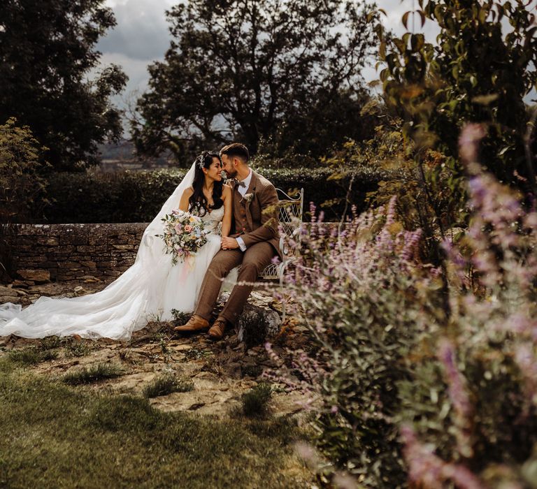 Bride in lace and tulle wedding dress and veil holding bright summer bridal bouquet sits with groom in brown tweed suit on bench in garden