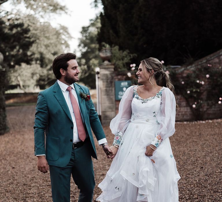 Groom in blue suit and pink tie holding hands with bride in long mesh sleeve wedding dress and white trainers walks across drive at Iscoyd Park