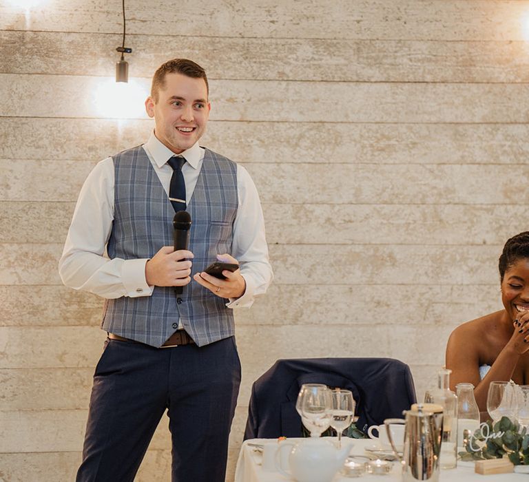 Bride smiles with Joy as the groom stands to do his speech on their wedding day