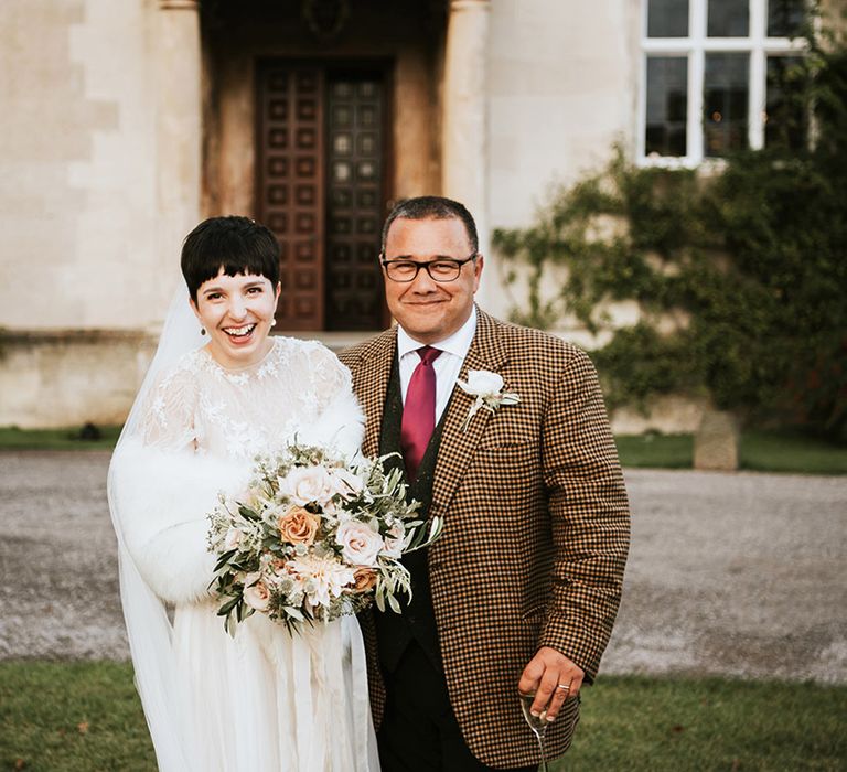 Bride with short hair in a white faux fur stole with her father in a check jacket outside Elmore Court 