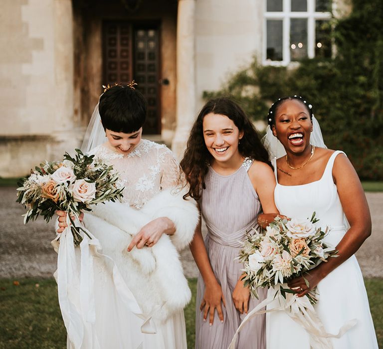 Two brides laughing with their junior bridesmaid in a dusky pink dress outside their country house wedding venue 