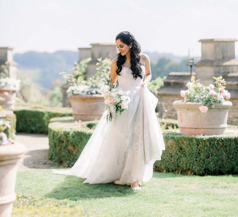 Indian bride holds her bridal gown outdoors on her wedding day whilst she holds floral bouquet 