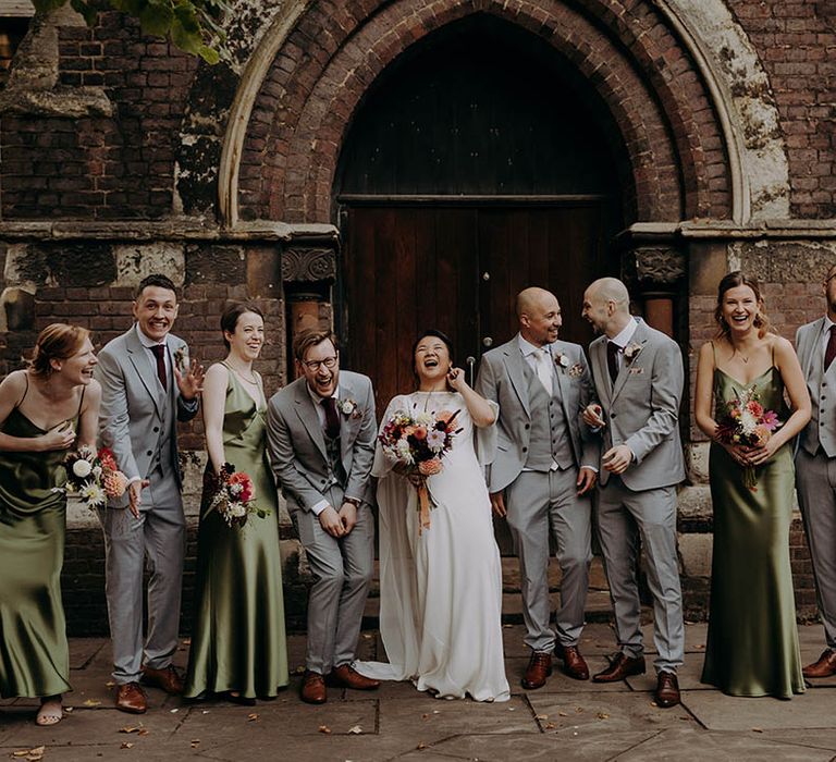 Bride & groom stand with their wedding party, as bridesmaids wear sleek satin olive green bridesmaids gowns and grooms wear grey three piece suits  | Irene Yap Photography