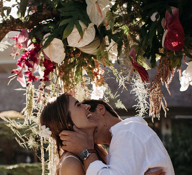 Groom kisses bride's neck under natural tropical flowers 