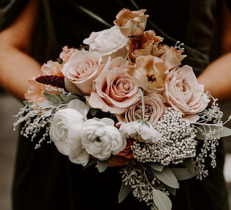 Bridesmaid in green velvet jumpsuit holds bouquet of roses and peonies and other flowers 