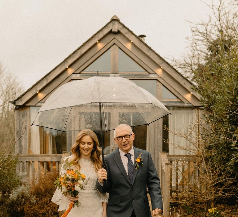 Father of the bride in blue suit holds umbrella as he walks the bride in lace a-line wedding dress to the ceremony 