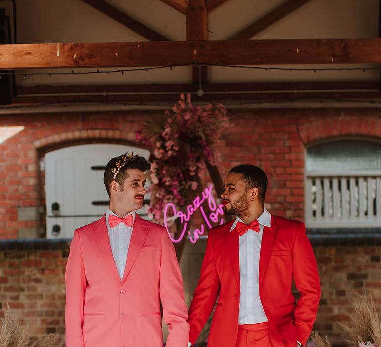 Groom in a red suit with winged eyeliner makeup holding hands with his partner in a pink suit, bow tie and gold crown