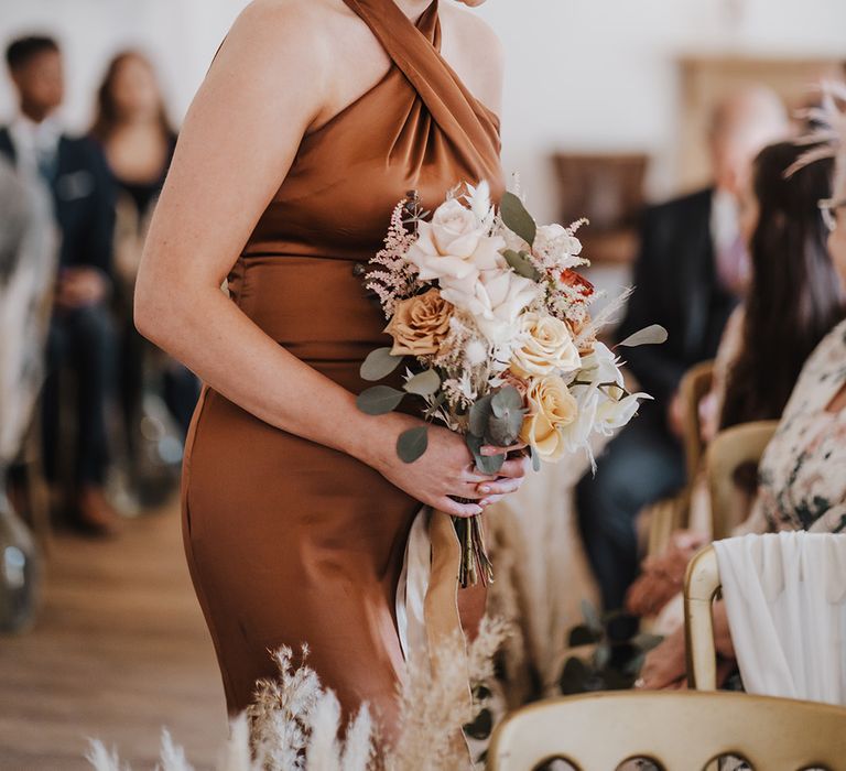 Bridesmaid in halter neck copper bridesmaid dress takes her seat for the wedding holding neutral wedding bouquet