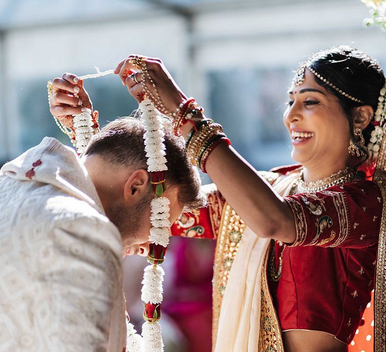 Bride in red traditional Indian wedding attire for multicultural wedding places a Jaimala on the groom