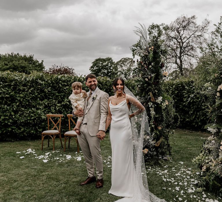 Bride in a minimalist slip wedding dress and bespoke pearl veil and groom in a beige suit at their outdoor garden wedding 