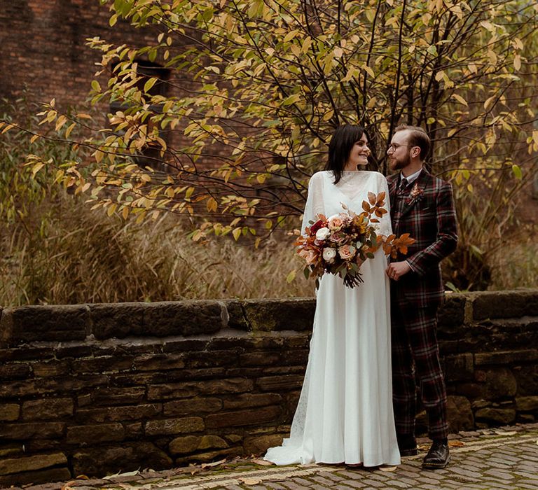 Groom stands behind bride on their wedding day as bride holds Autumnal inspired bouquet in front of trees outdoors 