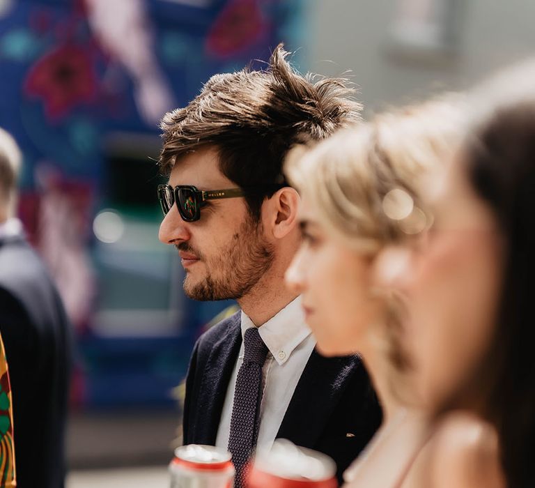 Wedding guests hold cans of beer as they stand outdoors in the sunshine 