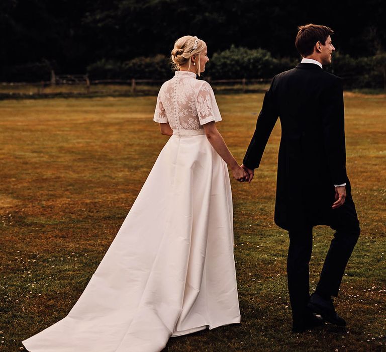 Bride in a lace button back wedding dress walks with the groom holding hands 